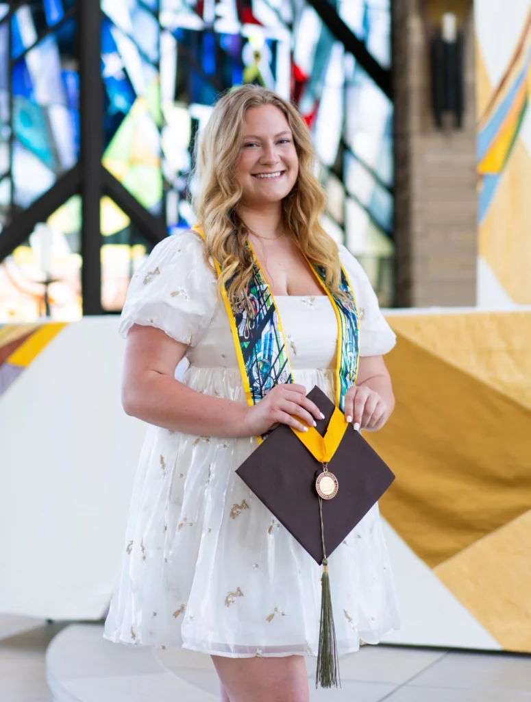 Smiling graduate Lauren Bartlett, M.S., in a white dress holding a cap, stands in front of colorful stained glass, embodying the spirit of a true Beacon.