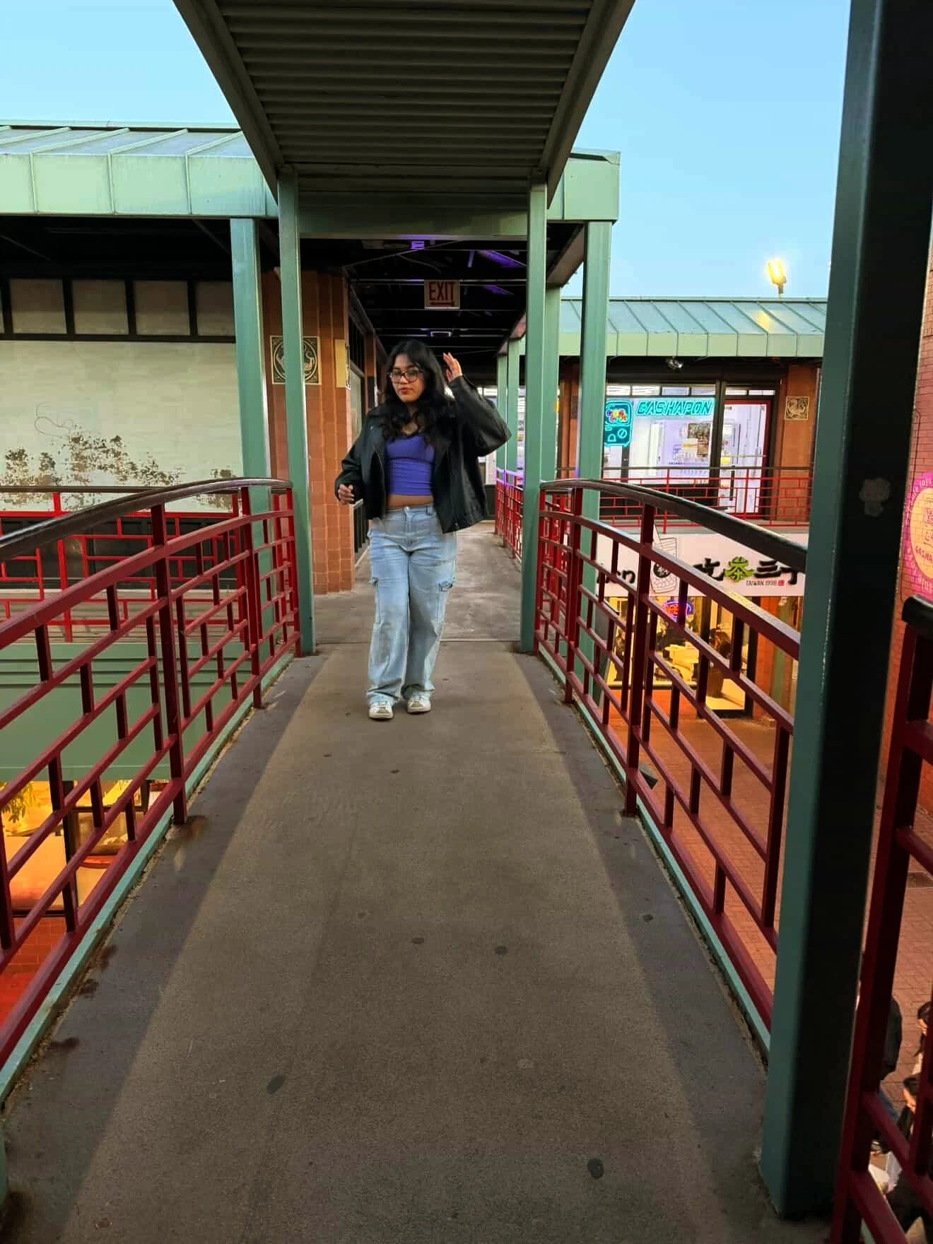 Ashley Sotelo, dressed in jeans and a jacket, walks on a bridge with red railings, smiling and waving.