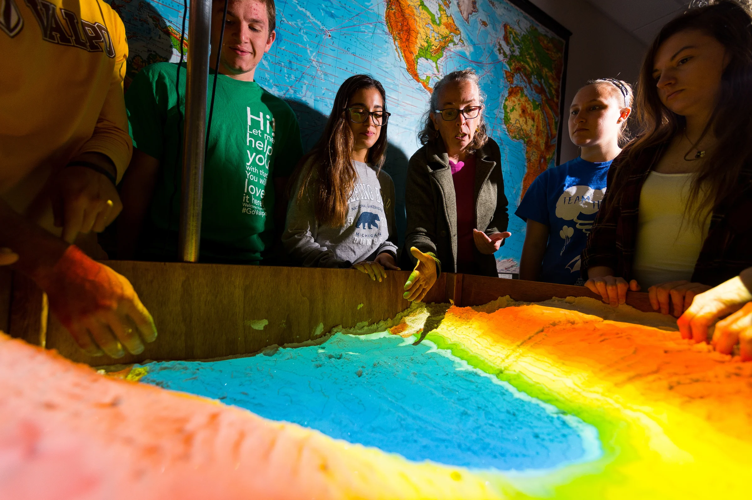Six people examine a colorful topographic sand table, commonly used in Environmental Science, with a world map displayed in the background.