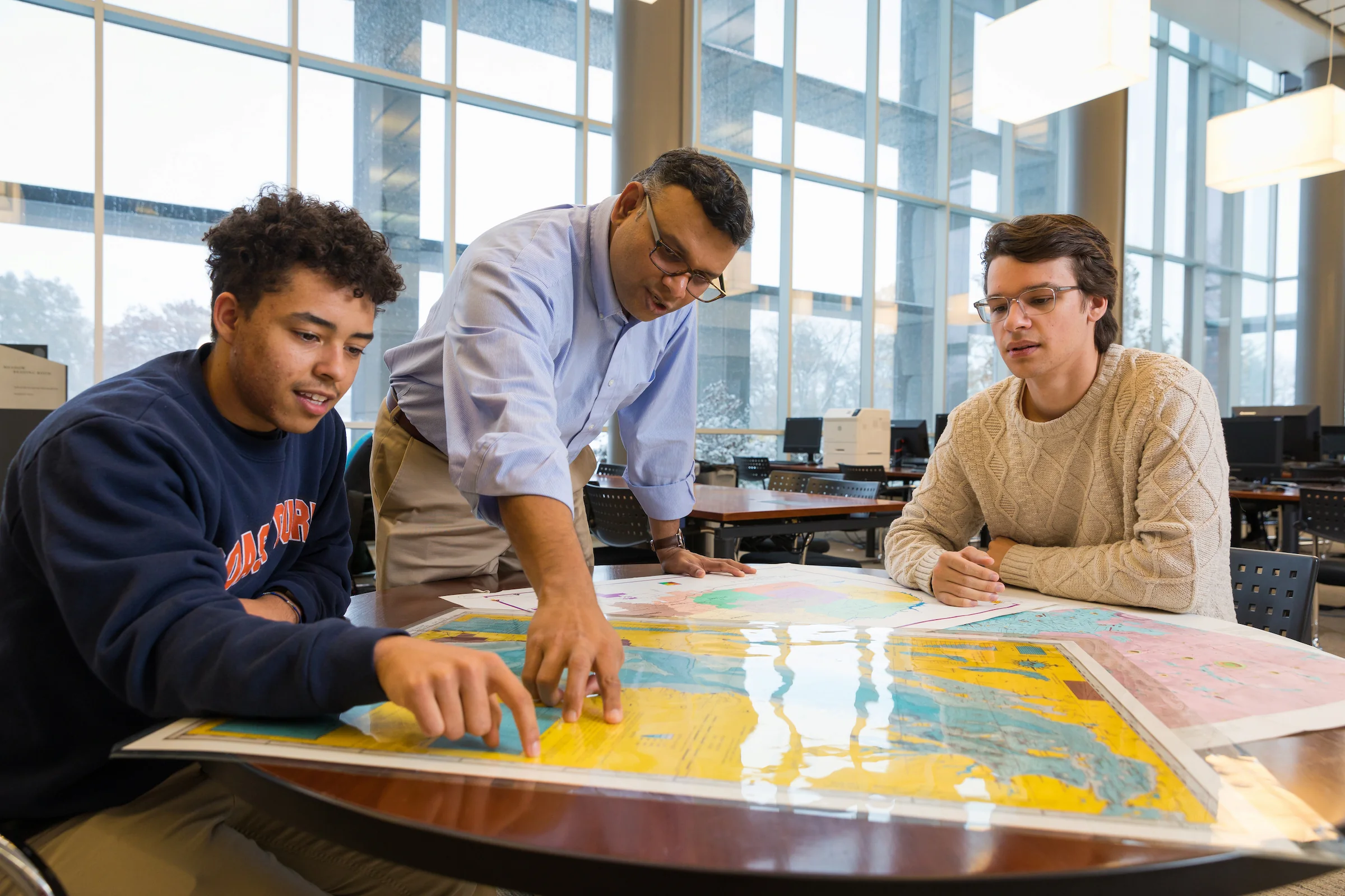 Three people sitting at a table study and discuss maps related to Environmental Science in a bright, modern room with large windows.
