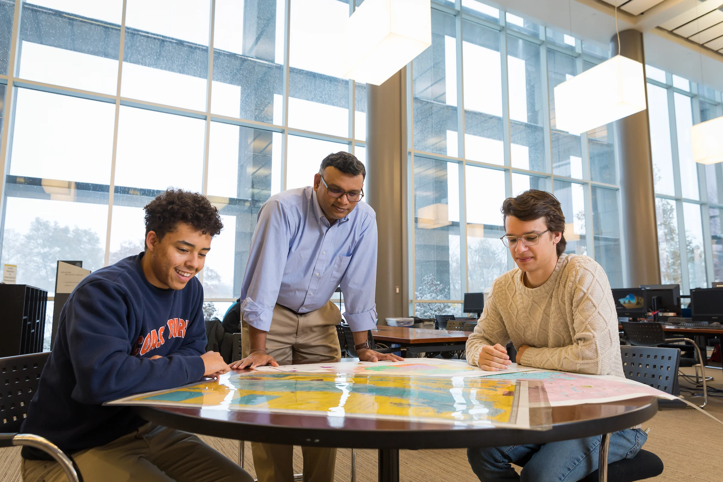 Three students from Valparaiso University look at a large map on a table in a bright, modern room with large windows, exploring concepts from Geospatial Sciences.