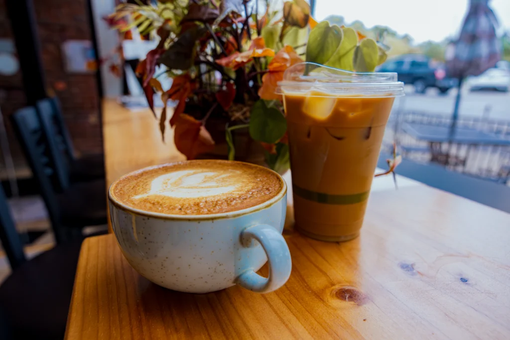 A cup of hot latte and a plastic cup of iced coffee sit on a wooden table near a plant, basking in the gentle morning light that streams through the window at Beacon, Walker Johnson and Ben Montgomery’s favorite local spot.