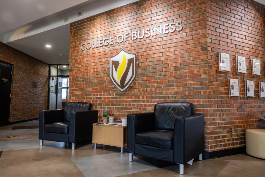 Two black chairs and a small table sit by a brick wall labeled “College of Business” with a shield logo, highlighting Valparaiso University’s impressive $1 Million Student-Run Investment Portfolio.