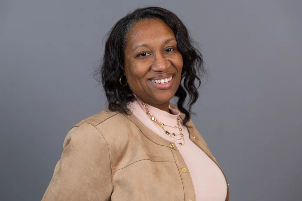 Smiling woman with wavy hair, wearing a tan jacket and pink top, poses in front of a gray background—perfectly capturing the spirit of CASS and its vibrant community of health professions students.