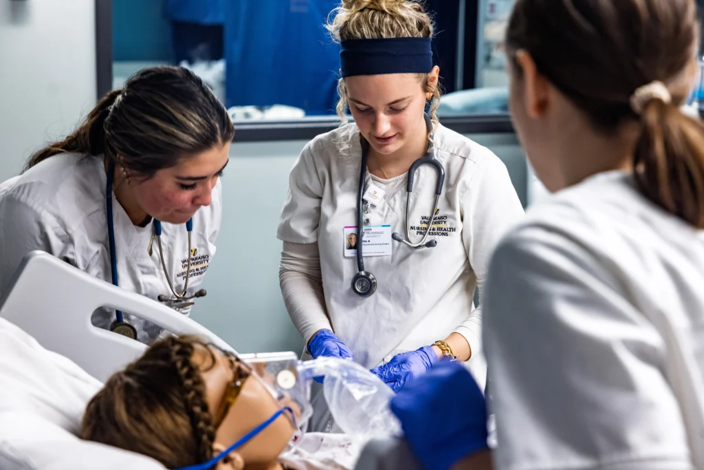 Three health professions students in white coats treat a patient in a hospital bed using medical equipment, demonstrating the importance of community care and collaborative learning.