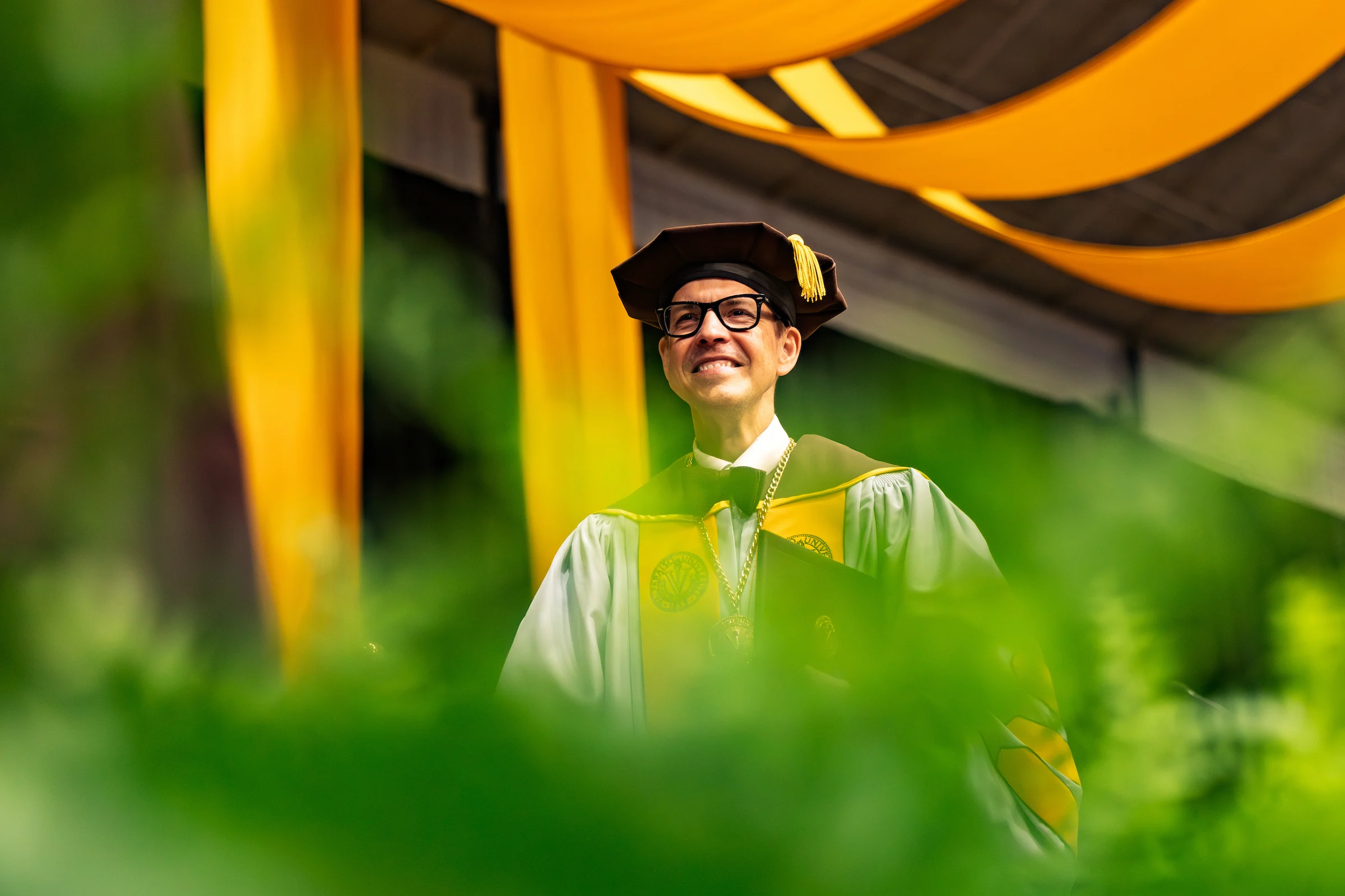 Rev. Brian Konkol in a cap and gown stands outdoors with yellow banners in the background.