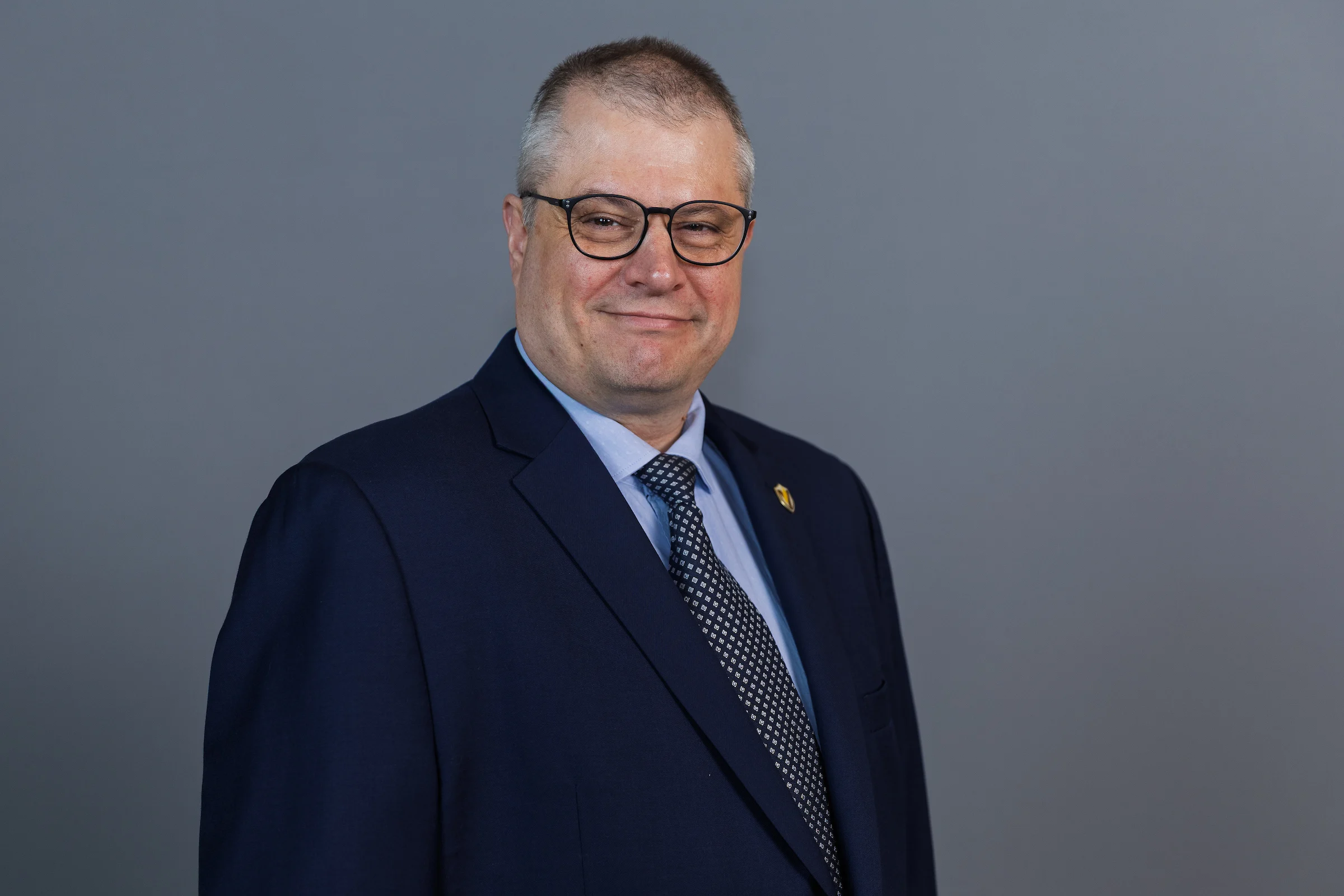 Man in a navy suit and glasses smiling, standing against a plain gray background. He is the Program Director for the Master of Organizational Leadership at Valparaiso University.