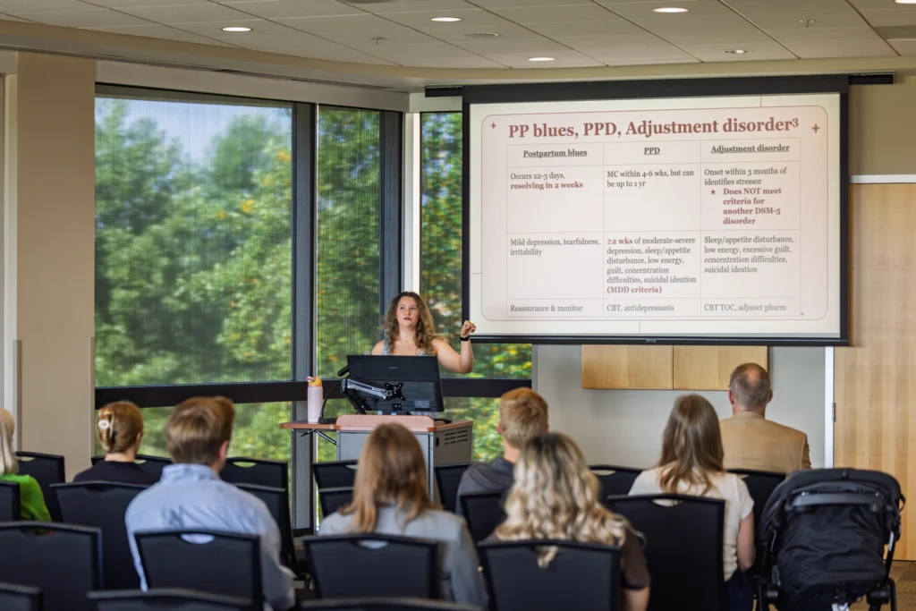 A woman gives a lecture on postpartum disorders to an audience of health professions students in a bright conference room, fostering awareness and community engagement.