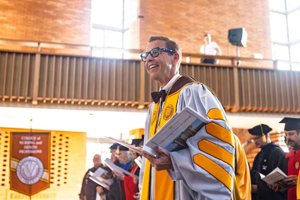 Smiling graduate in yellow and white gown holds program during Valpo’s Opening Convocation ceremony, celebrating with others and welcoming the Incoming Class.