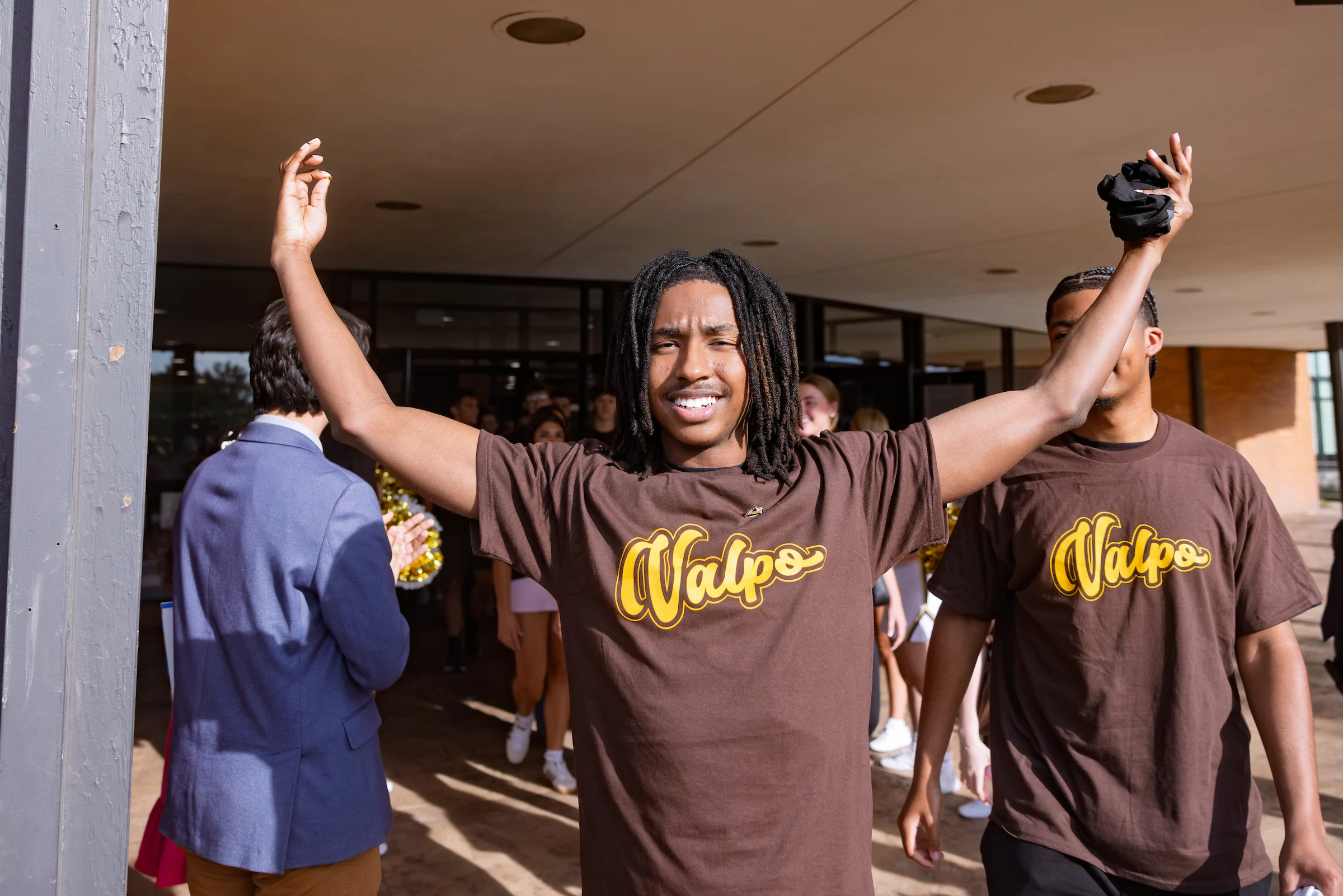 Smiling student in a “Valpo” shirt raises arms outside a building during Opening Convocation, with others from the incoming class in similar shirts nearby.