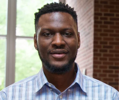 Man with a beard wearing a blue plaid shirt, standing indoors by a window and brick wall, Dr. Chinedu Ufodike, Ph.D., exudes a thoughtful and scholarly presence.