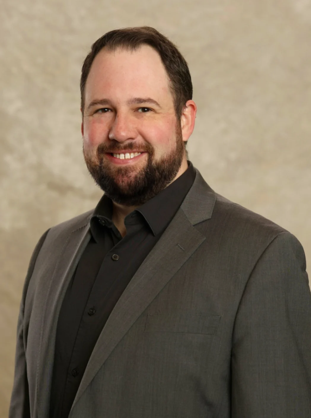 Man with a beard smiling, wearing a gray suit jacket and black shirt, in front of a beige background—Jordan Dollins exudes confidence and style.