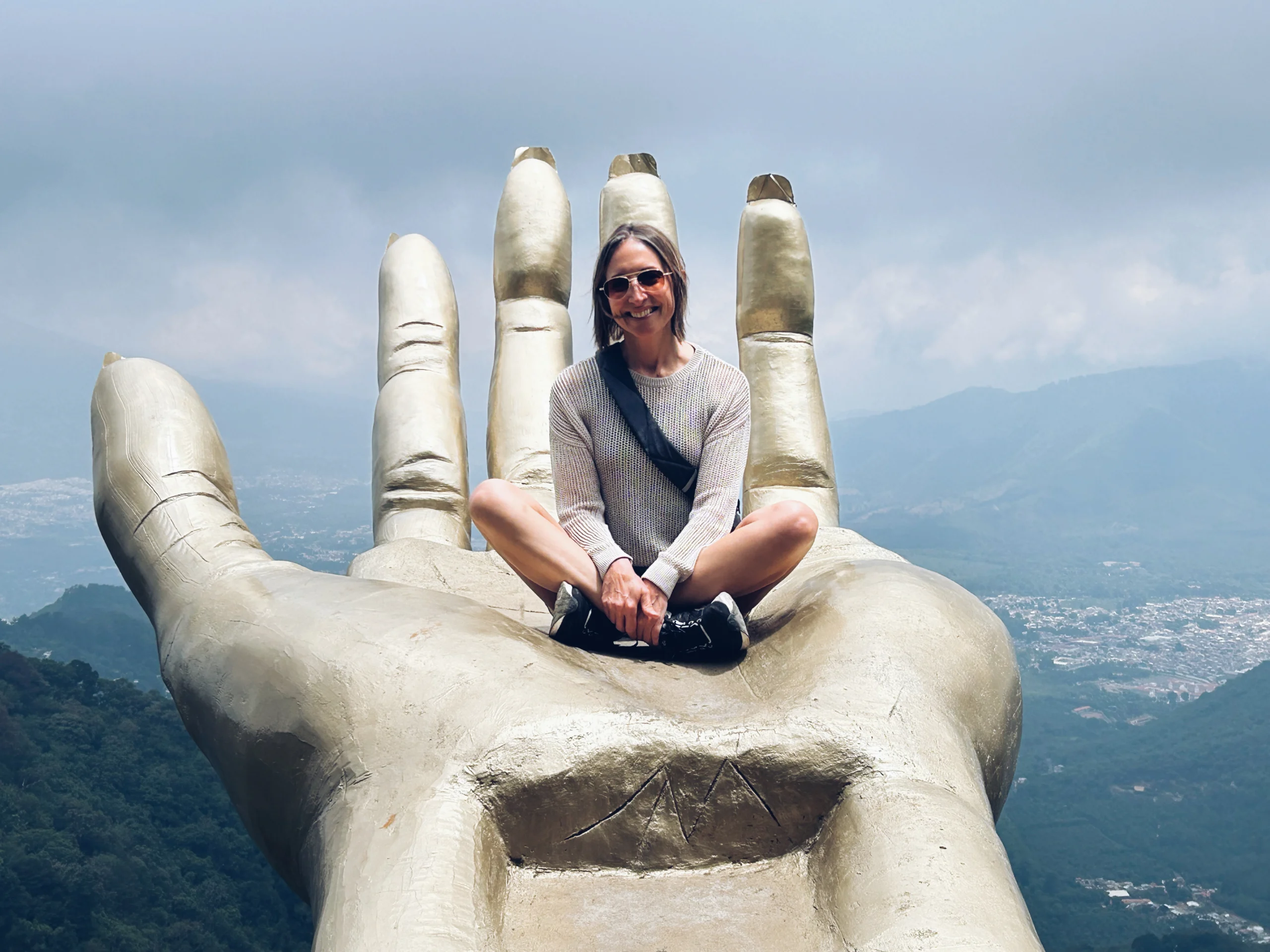 A woman sits cross-legged on a giant hand statue with mountains and sky in the background, embodying the spirit of Valparaiso University’s College of Nursing and Health Professions.