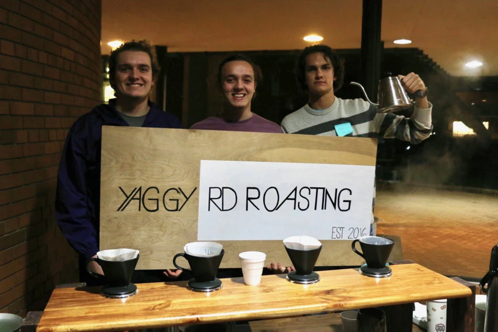 Three people—Ben Montgomery, Walker Johnson, and a third team member—stand behind a coffee stand holding a Yaggy Rd Roasting sign; Beacon-branded coffee items are neatly arranged on the table.
