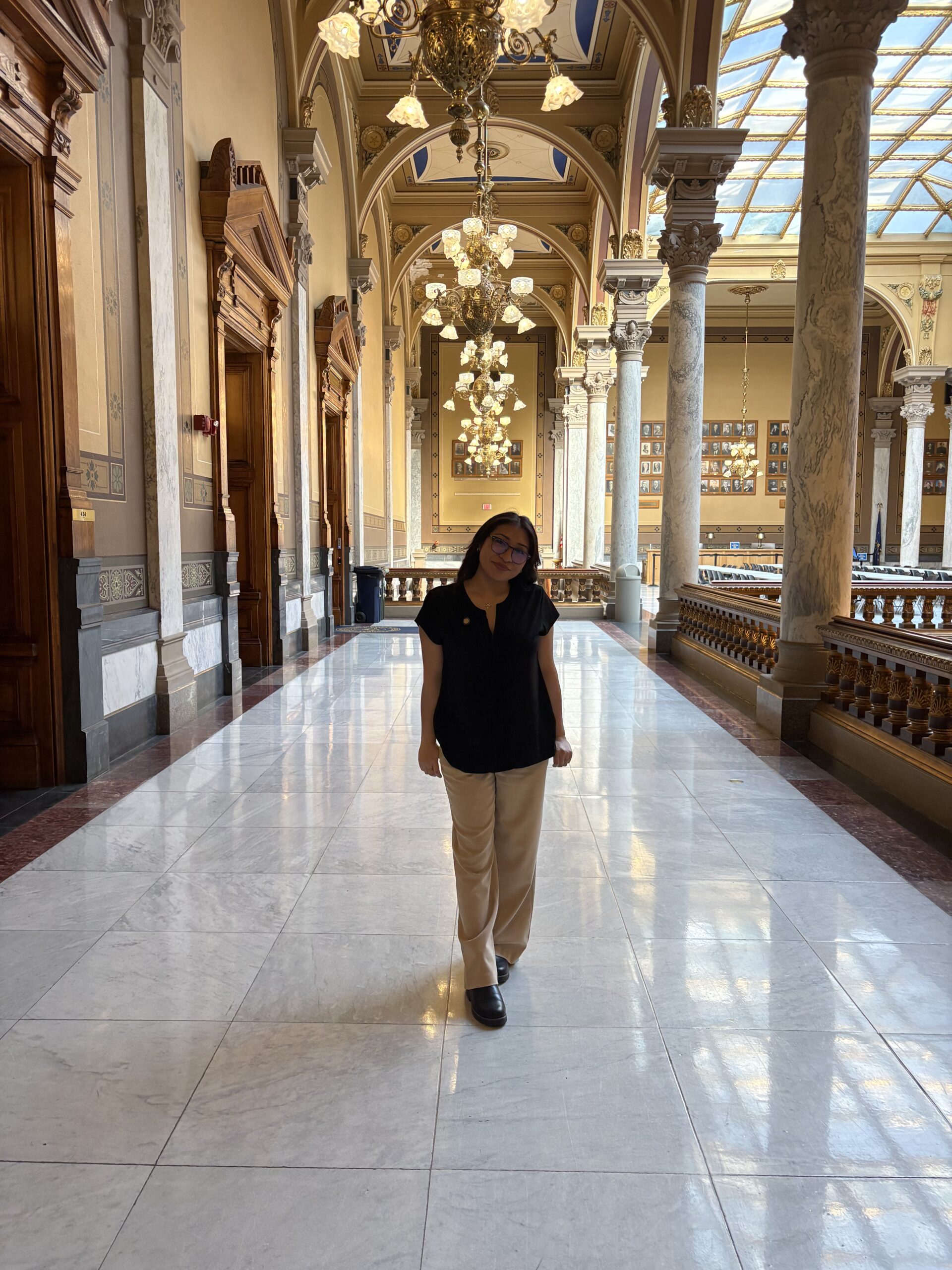 Person in black shirt and beige pants stands in a grand hall with columns, chandeliers, and a glass ceiling, reminiscent of the distinguished settings often attended by UPC President Daniela Martinez.