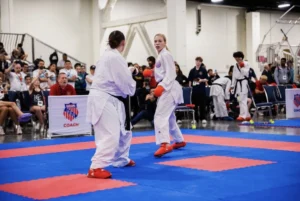 Two karate athletes spar in a competition, watched by an audience and officials in a gymnasium setting, as Valpo’s own World Champion Hailey Reichelt showcases her impressive skills.