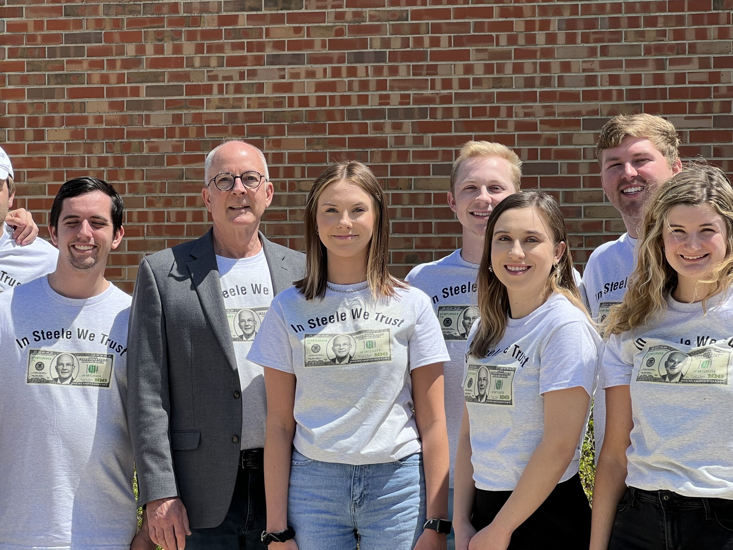Group of people in matching “In Steele We Trust” shirts standing in front of a brick wall, smiling at the camera—members of Valparaiso University’s renowned Student-Run Investment Portfolio managing over $1 Million.
