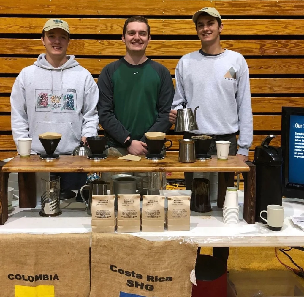 Three smiling people—Ben Montgomery, Walker Johnson, and a colleague—stand behind the Beacon coffee booth with brewing equipment, bags of coffee, and cups.
