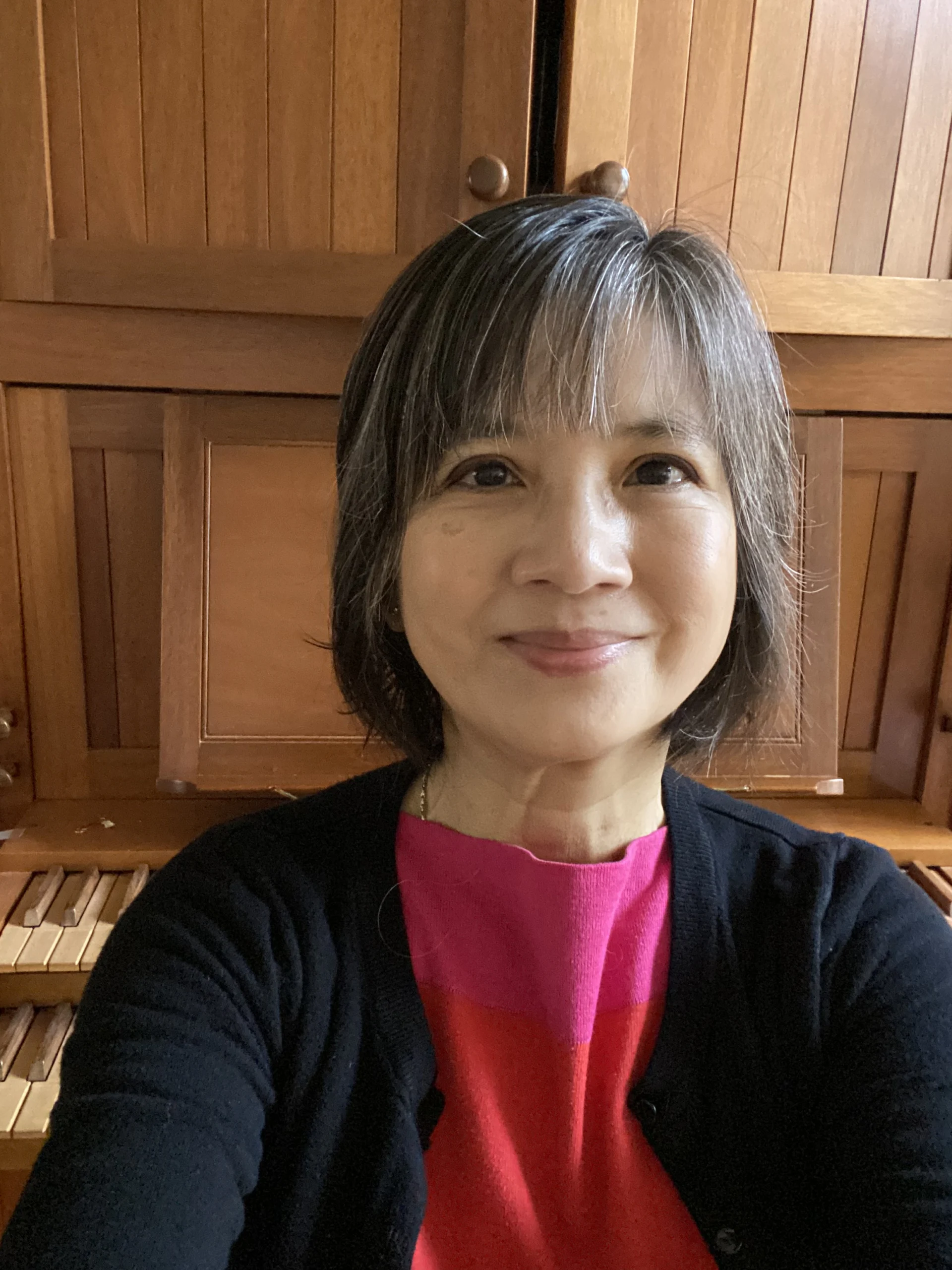 Szu-Ping Chang, a woman with short, gray-streaked hair, smiles warmly while wearing a pink top and black sweater in front of a wooden organ.