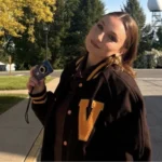 Person in a black varsity jacket, featuring a large “V,” holds a camera and smiles outdoors with trees in the background, embodying the welcoming spirit of the LINC Language and Intercultural Learning Center.
