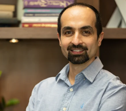 Smiling man with short hair and beard in a light blue shirt standing in front of a bookcase—this is Mohammad Fathi, Ph.D.