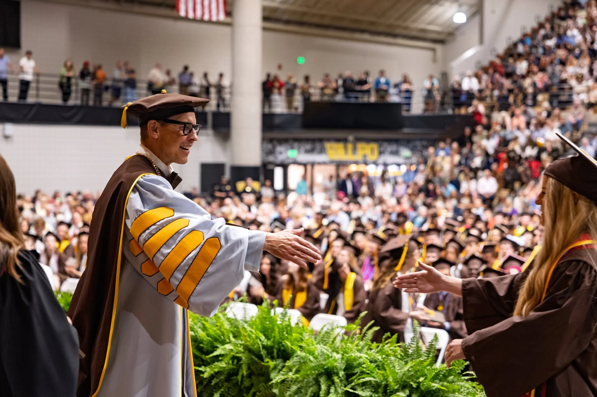 A person in academic regalia representing the President’s Office reaches to shake hands with a graduate at a crowded commencement ceremony.