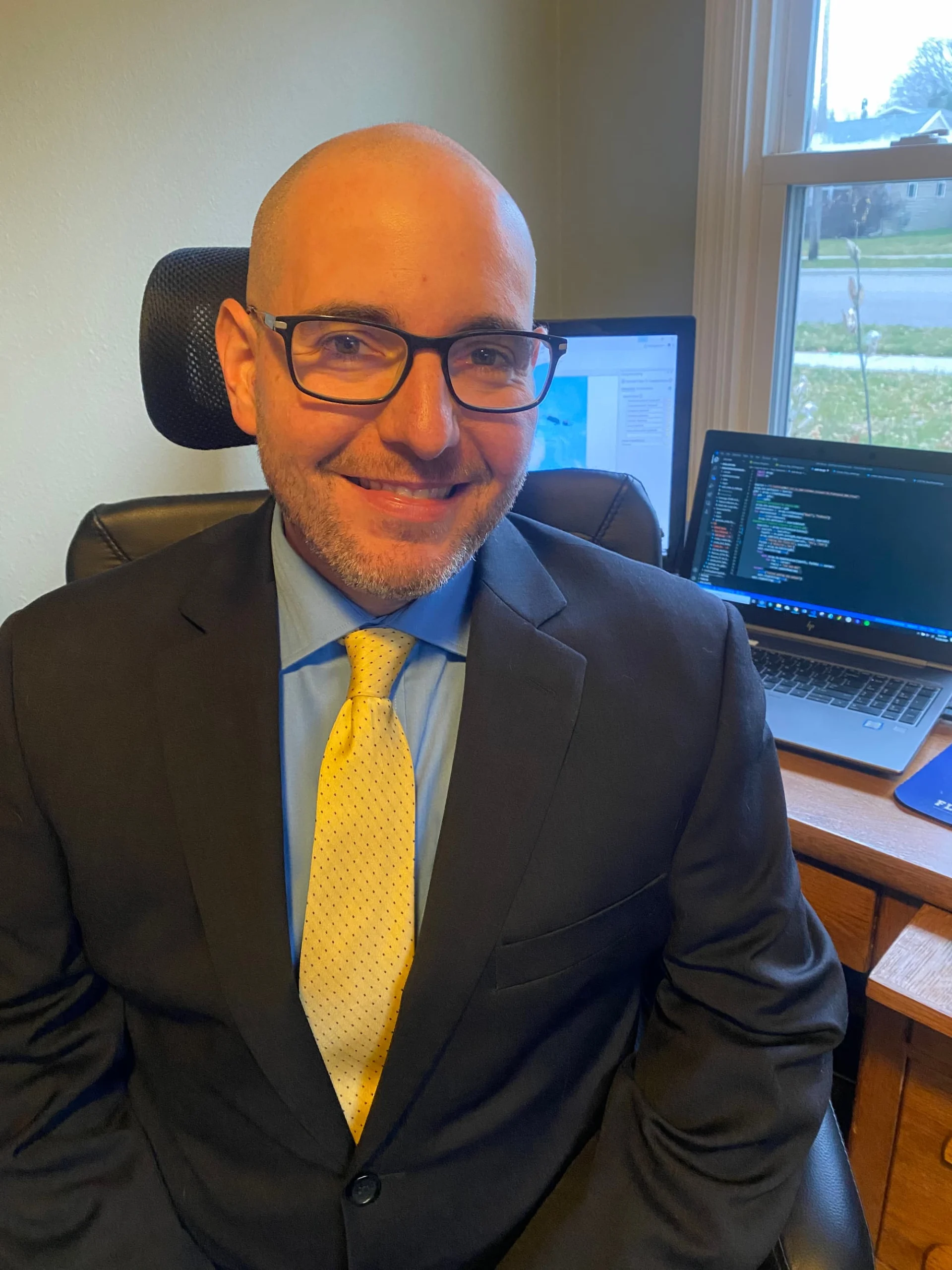 Smiling Zach Hassler, dressed in a suit and yellow tie, sits at a desk with computers in a home office.
