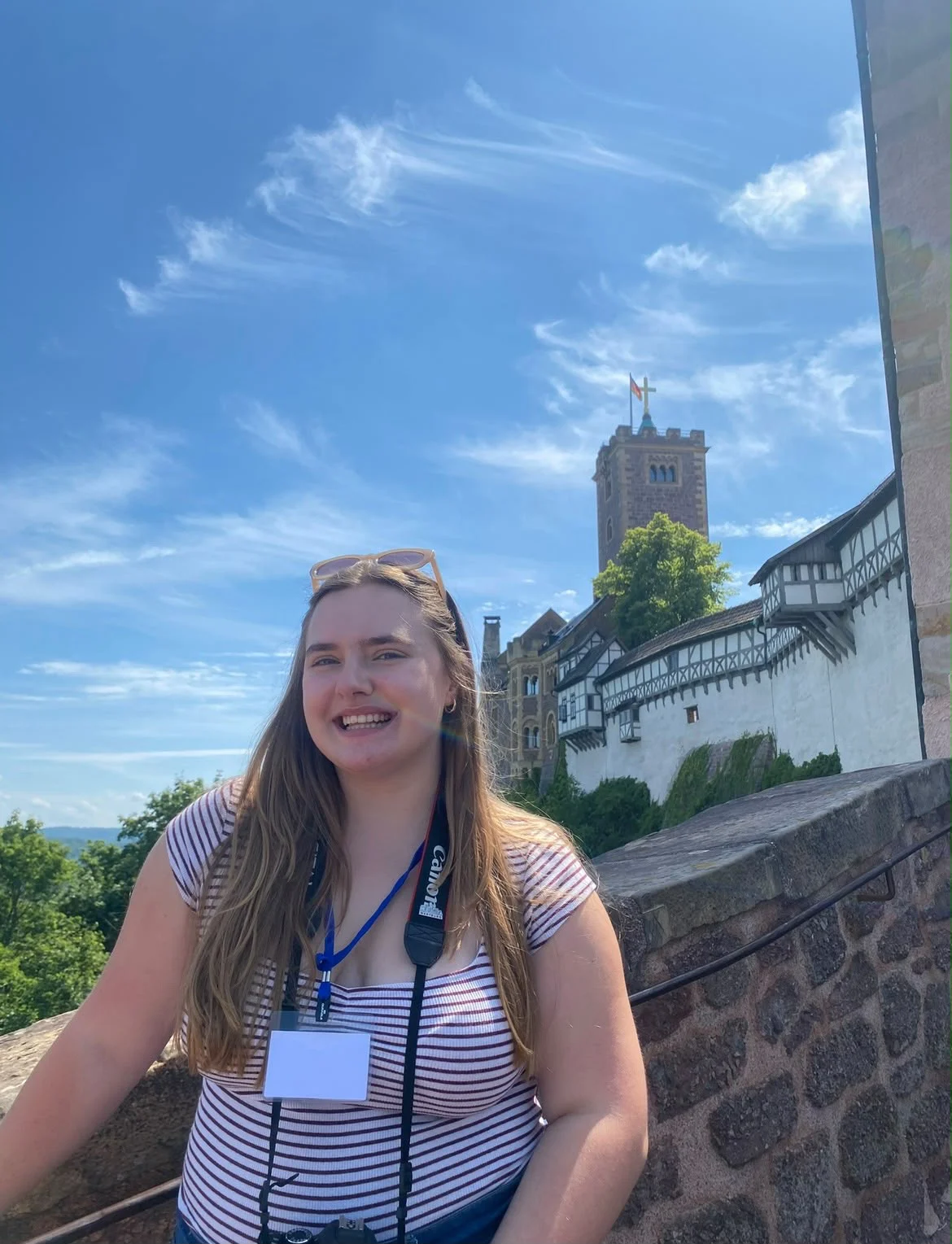 Smiling woman with a camera stands by a stone wall, with a castle and blue sky in the background—Nathalia Lenz Prochnow, UPC Vice President, enjoying a picturesque day out.