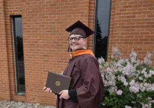 Storm Pendleton, dressed in a maroon graduation gown and cap, holds a diploma while smiling outside near flowers and a brick building—ready to make an impact in healthcare leadership and champion community well-being.