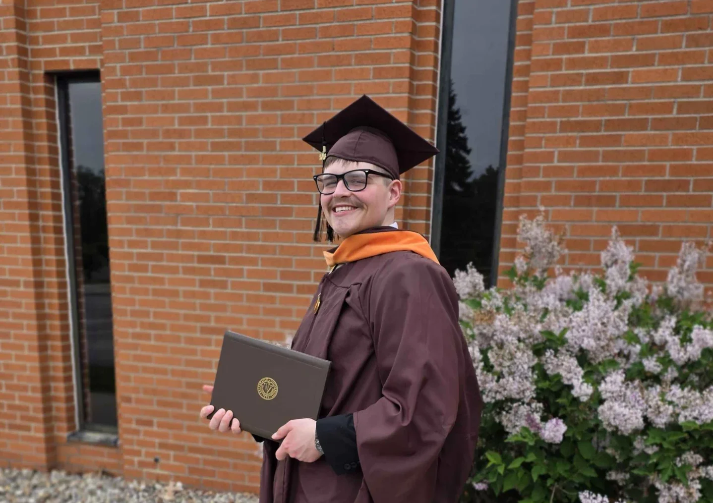 Storm Pendleton, dressed in a maroon graduation gown and cap, holds a diploma while smiling outside near flowers and a brick building—ready to make an impact in healthcare leadership and champion community well-being.