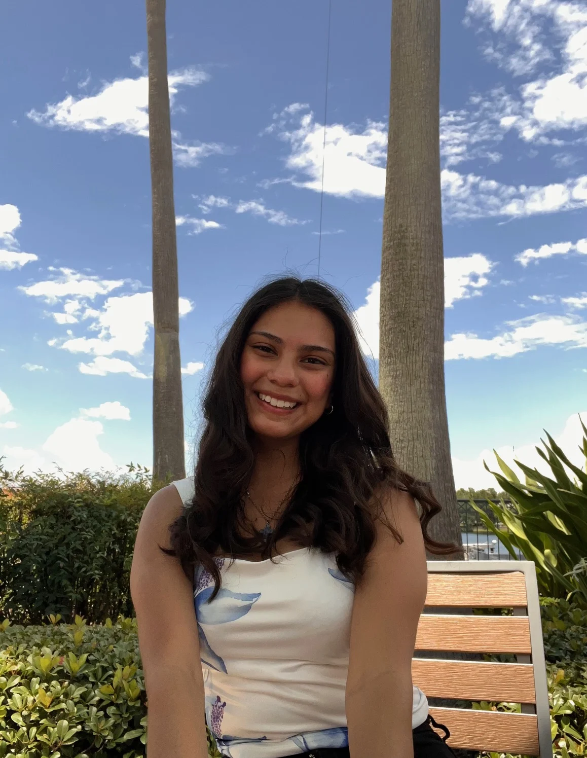 Young woman with long hair smiles while sitting on a bench outside, with blue sky and palm trees behind her, capturing a candid moment reminiscent of Ashley Sianez’s carefree spirit.