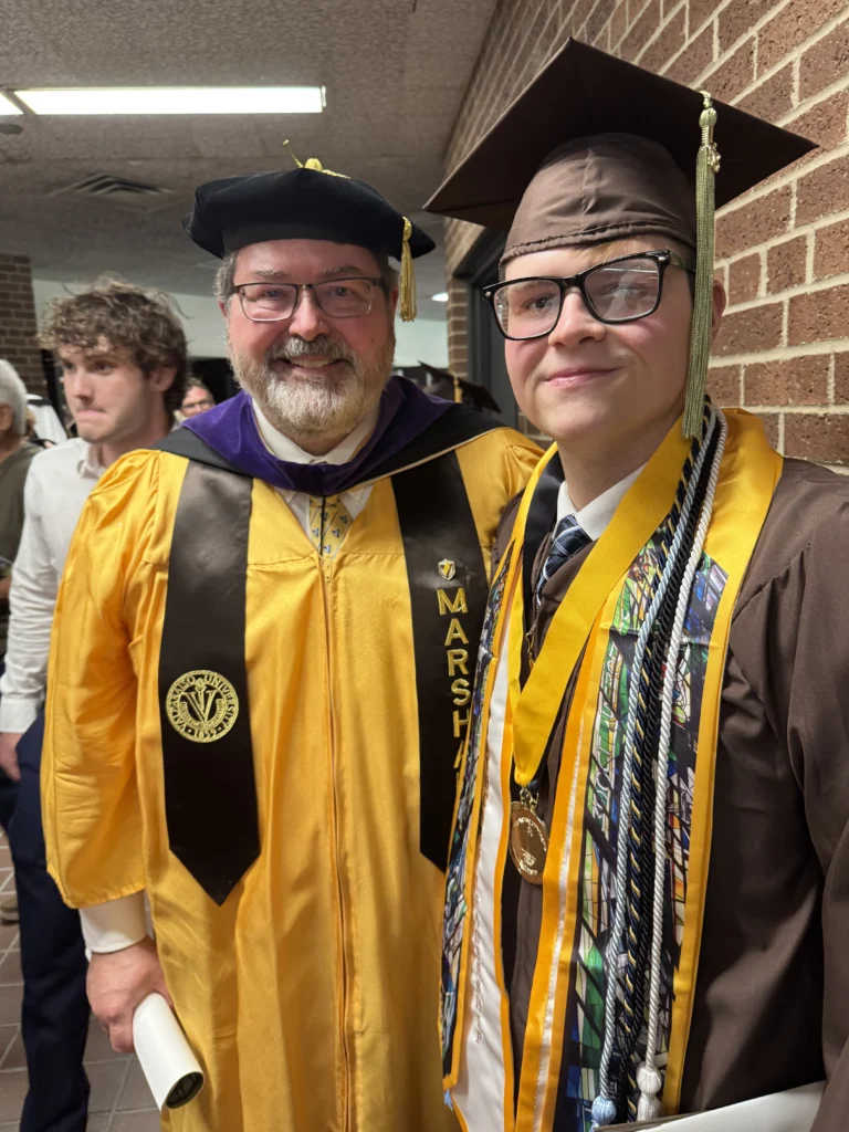 Two people in gold graduation gowns and caps stand smiling indoors at a graduation ceremony, celebrating their achievements in healthcare leadership and commitment to community well-being.