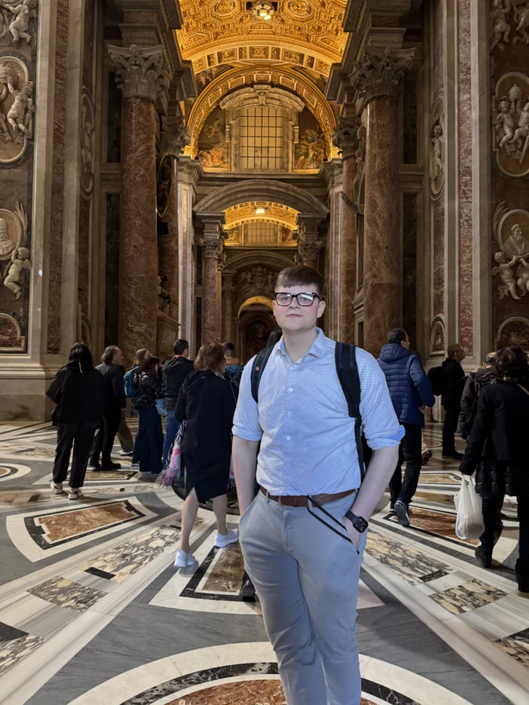 Man in a white shirt stands in an ornate hall with marble columns and a golden, decorated ceiling, embodying the spirit of healthcare leadership dedicated to advancing community well-being.