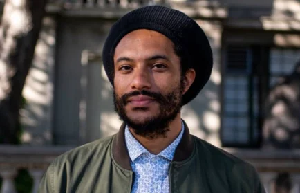 A man with a beard and black hat stands outside, wearing a green jacket and patterned shirt, smiling slightly—as if on his way to attend a Public Lecture by Jovan Scott Lewis on Spatial Practice.