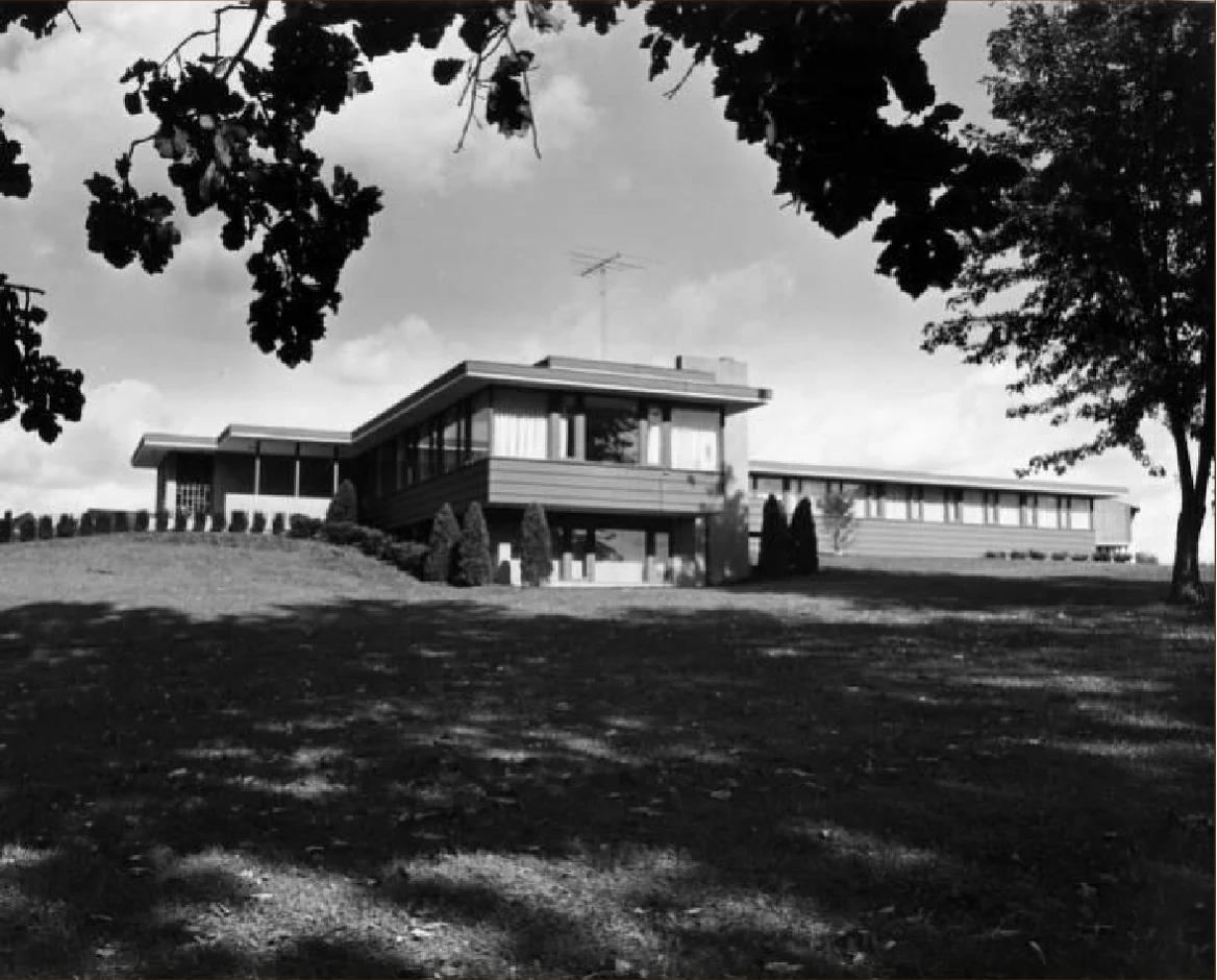 Black-and-white photo of a modern, flat-roofed building viewed from a grassy lawn with tree branches above, capturing a moment where contemporary design meets history in a single frame.