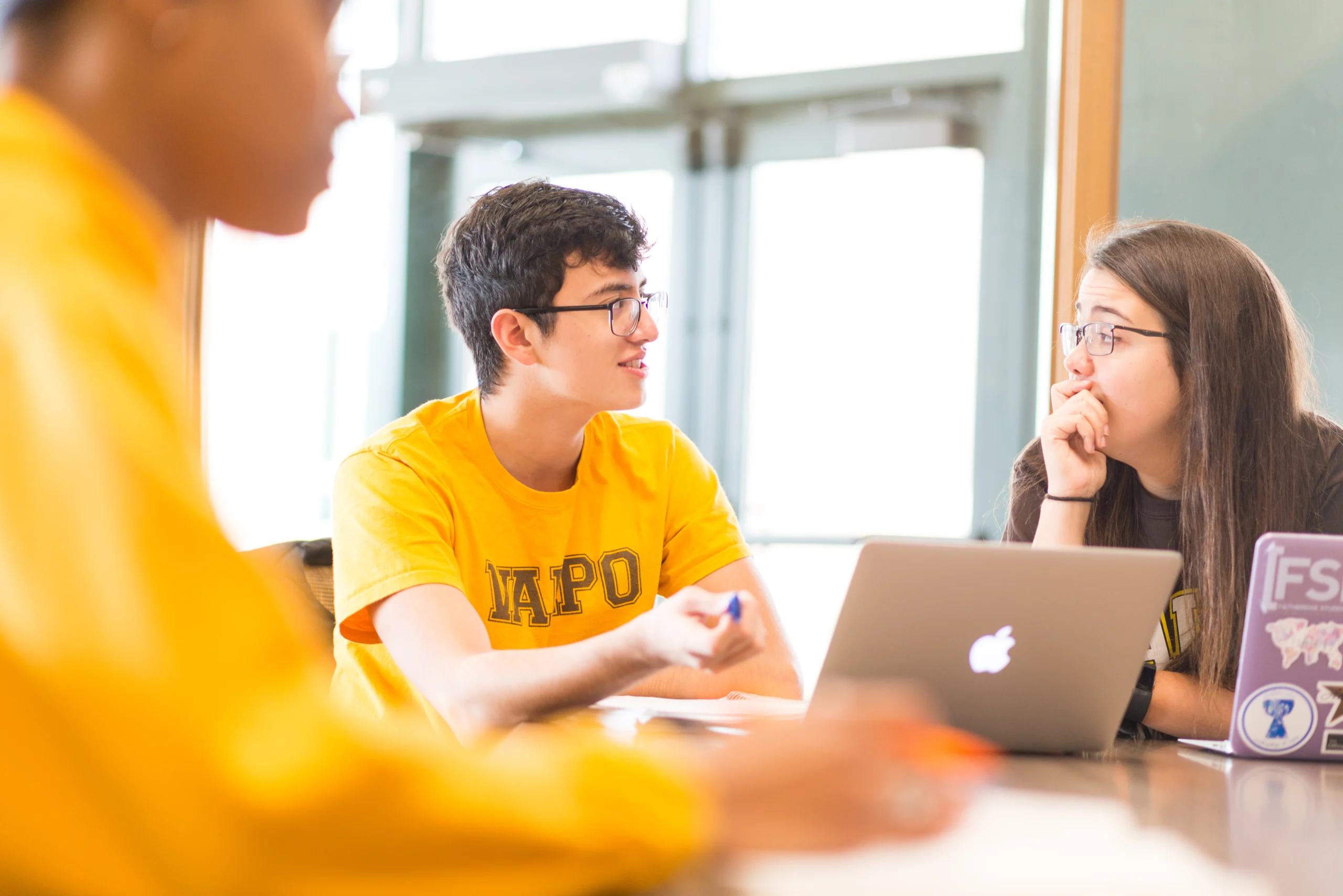 Three students in yellow shirts talk around a table with a laptop in a brightly lit room, sharing insights about what to expect during the first semester of grad school.