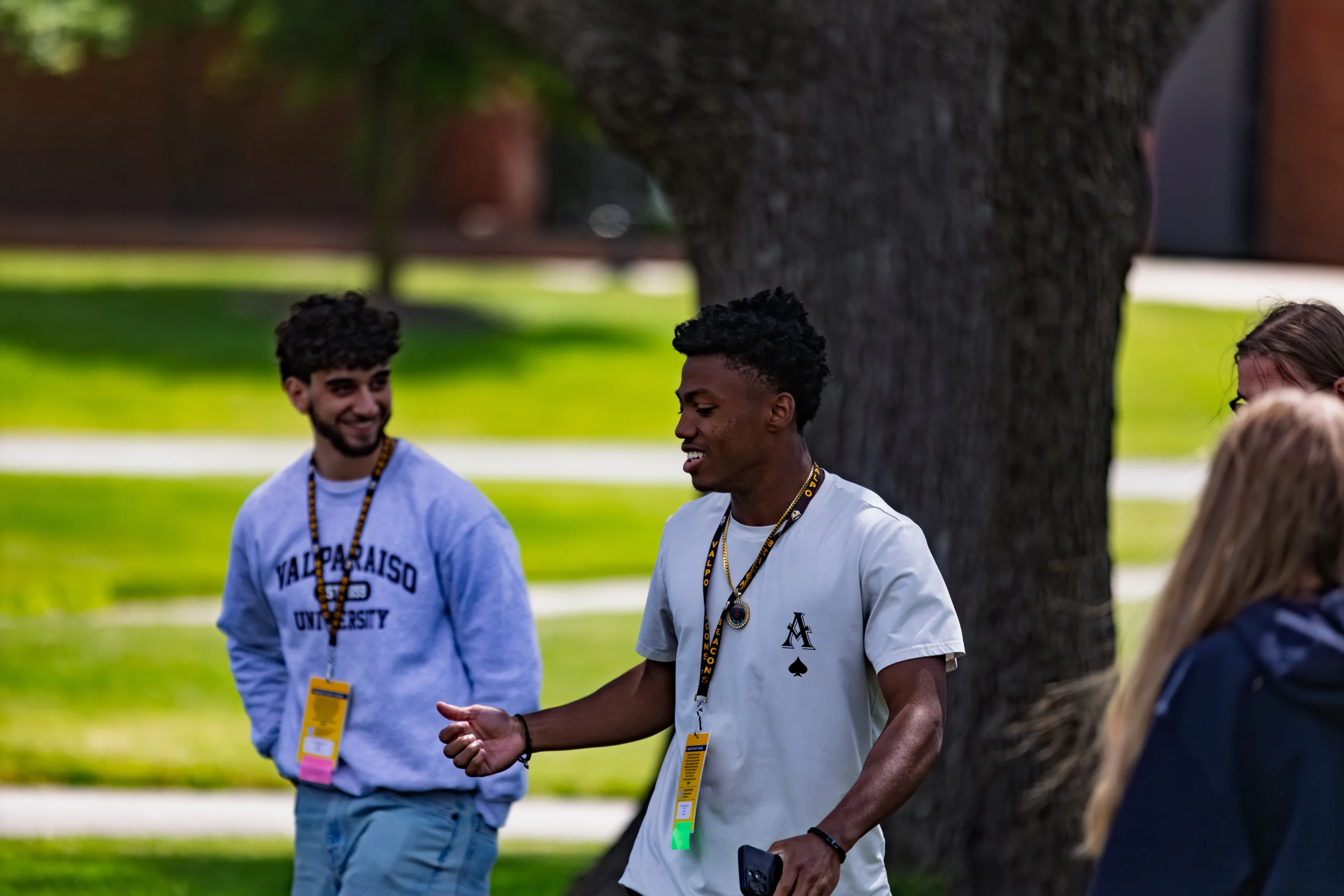 Two young men with name tags walk and talk outside, smiling, with trees and grass in the background, as they share college social tips on how to adjust socially during their first semester of college.