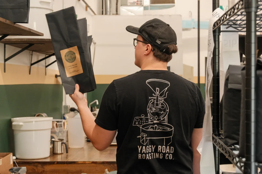 A person in a cap and branded shirt holds a large black coffee bag inside the Beacon roasting facility.