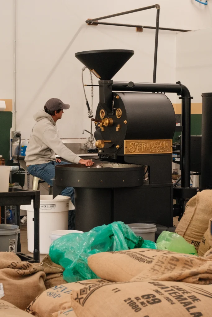 A person operates a large coffee roasting machine in a workspace filled with coffee bags, embodying the dedication to craft found at Beacon, where artisans like Ben Montgomery and Walker Johnson have honed their expertise.