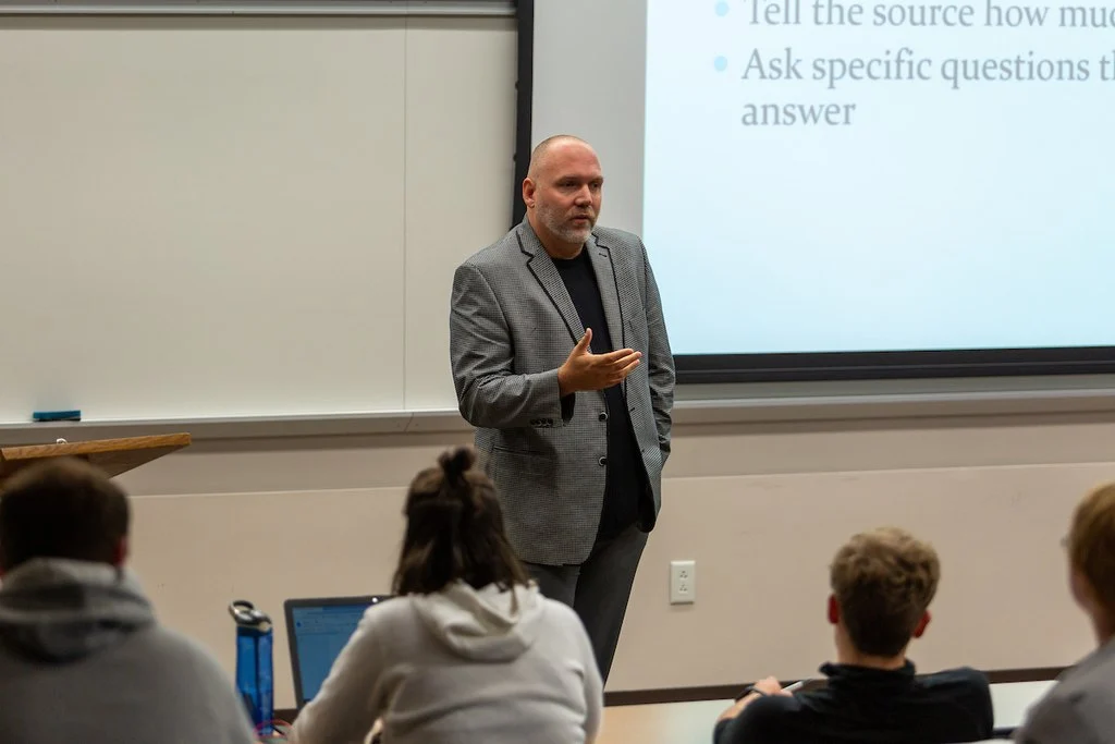 A man in a gray blazer, identified as Paul Oren, speaks to students in a Valparaiso classroom with a projected presentation behind him, possibly related to his work with The Beacon.