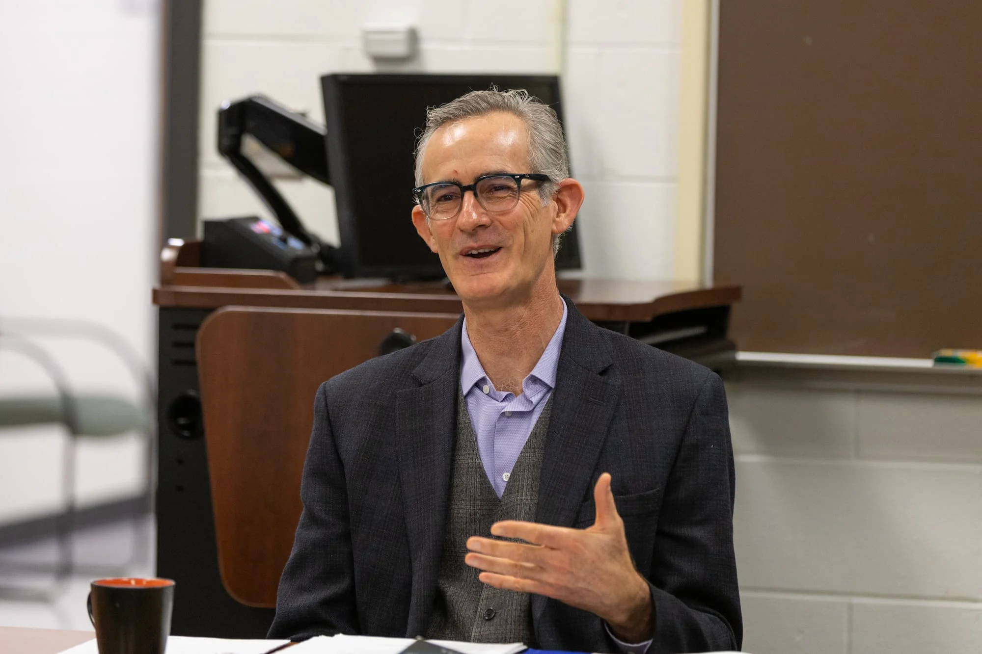 A man in glasses and a suit gestures while speaking about ethics in an office or classroom setting.