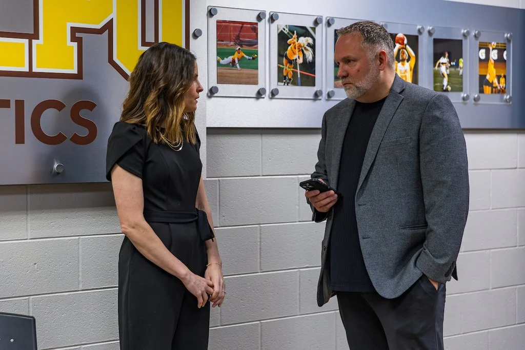 Two people converse indoors near a wall with sports photos; one holds a phone, both look serious, perhaps discussing an article from the Valparaiso University Beacon or insights shared by Paul Oren.