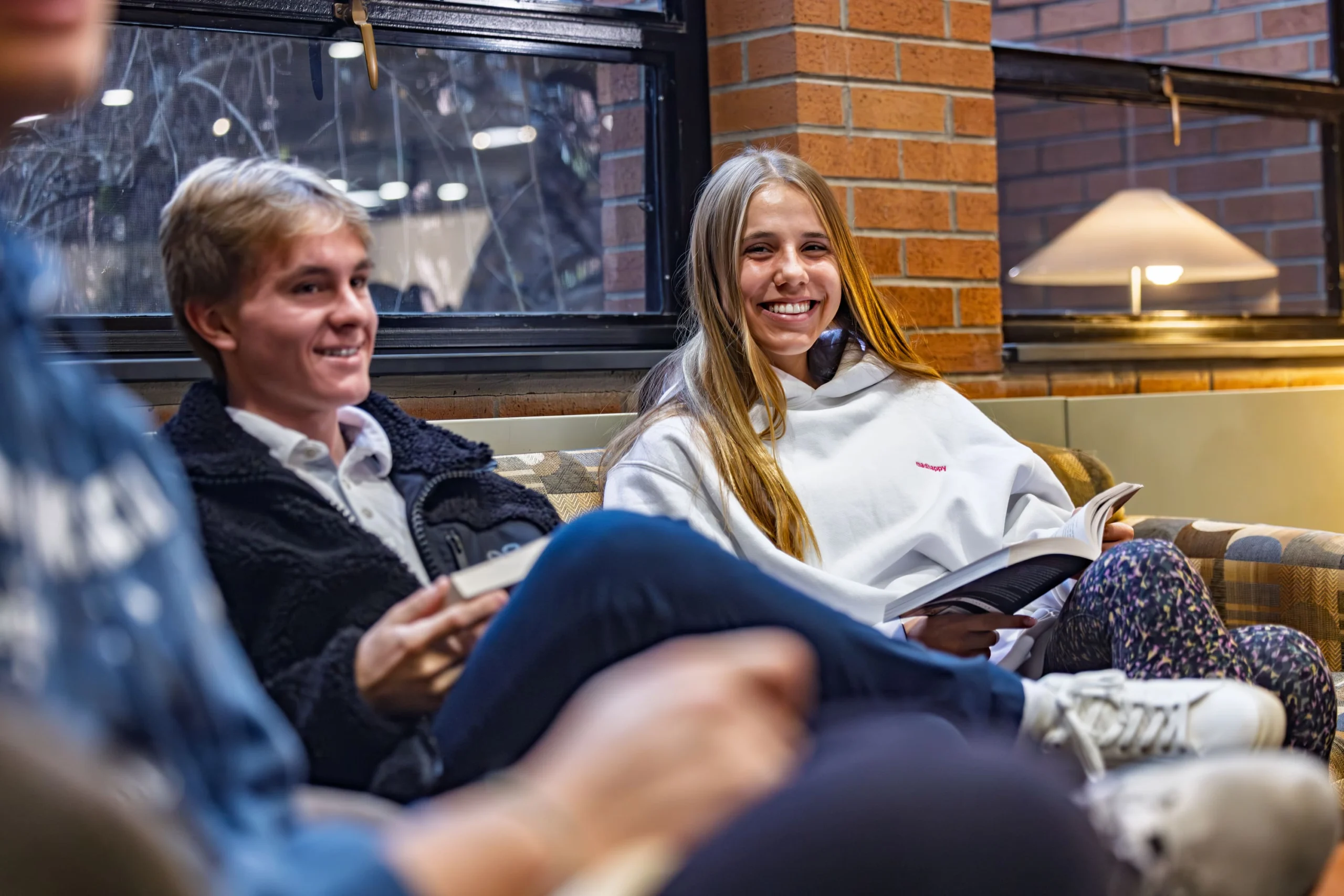 Two young people sit on a couch indoors, smiling and holding books about ethics and society, with another person in the foreground.