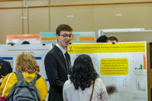 A man in a suit discusses a tornado warning research poster with two women at a science fair, emphasizing the importance of public safety and highlighting recent studies conducted at Valparaiso University.