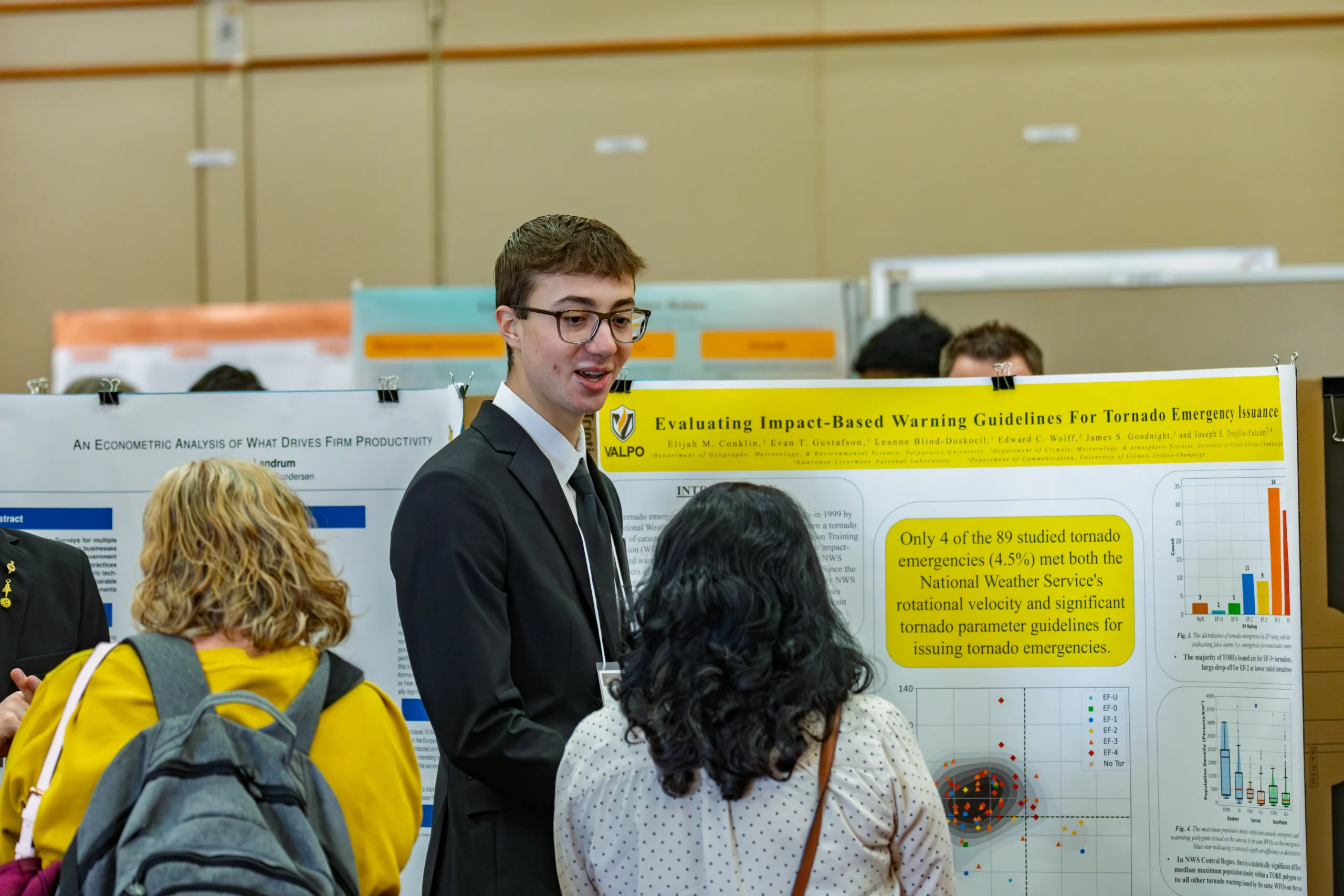 A man in a suit discusses a tornado warning research poster with two women at a science fair, emphasizing the importance of public safety and highlighting recent studies conducted at Valparaiso University.