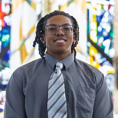 A Valparaiso University student in a gray shirt and striped tie stands in the Chapel of the Resurrection, with stained glass in the background.