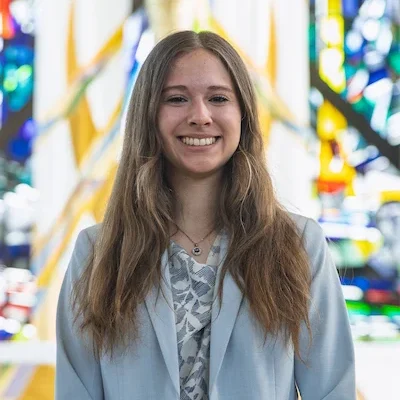 Smiling Valparaiso University student with long brown hair wears a light blue blazer, standing in front of stained glass windows in the Chapel of the Resurrection on campus.