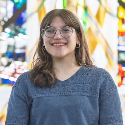 A Valparaiso University student with glasses and a blue sweater smiles, standing in front of a colorful stained glass window in the Chapel of the Resurrection on campus.