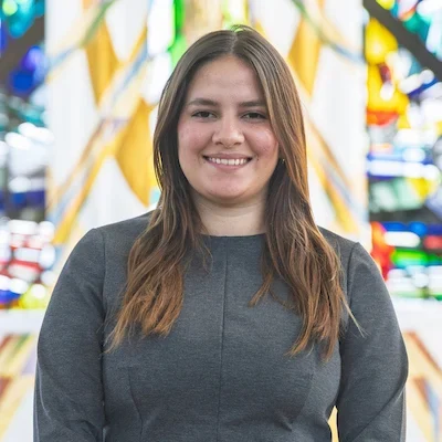 Valparaiso University student with long brown hair in a gray top smiles, standing in front of colorful stained glass in the Chapel of the Resurrection on campus.