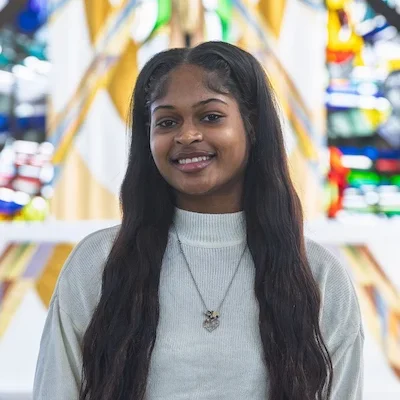 Smiling Valparaiso University student with long hair, wearing a white sweater, stands in the Chapel of the Resurrection with colorful stained glass behind her.